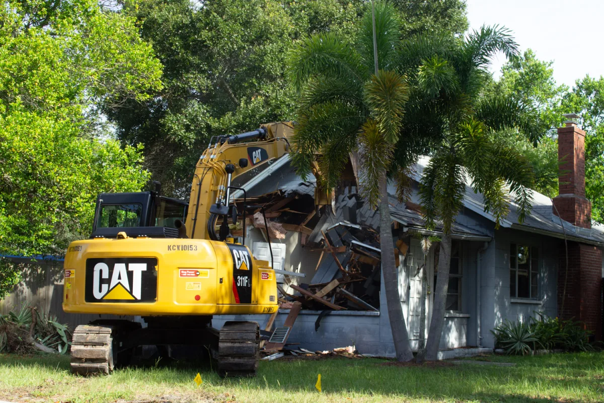 An excavator with a grapple attachment tearing down a single-story wood-frame house, with broken framing and debris piled around the machine.